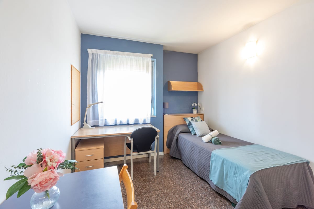 Una habitación pequeña y luminosa, con escritorio, silla y cama con una manta azul.