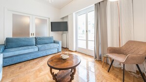 Una sala de estar con un sofá azul, una mesa de centro de madera, un sillón marrón, un televisor de pantalla plana y un ventanal con cortinas.
