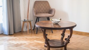 Una mesa redonda de madera con una cesta tejida encima, una silla y un taburete pequeño en una habitación con suelo de parqué de madera y cortinas de color claro.