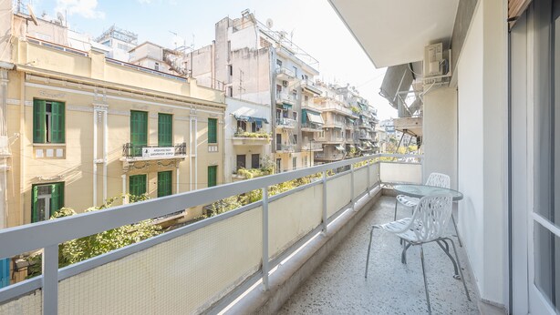 Un balcón con una mesa y sillas, con vista a una hilera de edificios con balcones y contraventanas verdes.