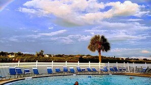 A swimming pool with lounge chairs and a palm tree, under a partly cloudy sky with a rainbow.