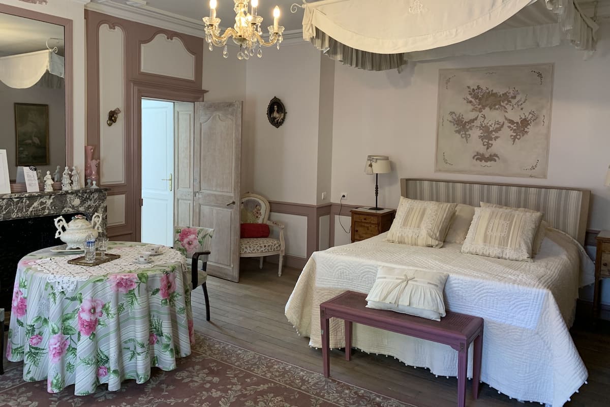 Un dormitorio con una cama grande, una mesita de noche, una silla, una pequeña mesa con mantel de flores y un candelabro.