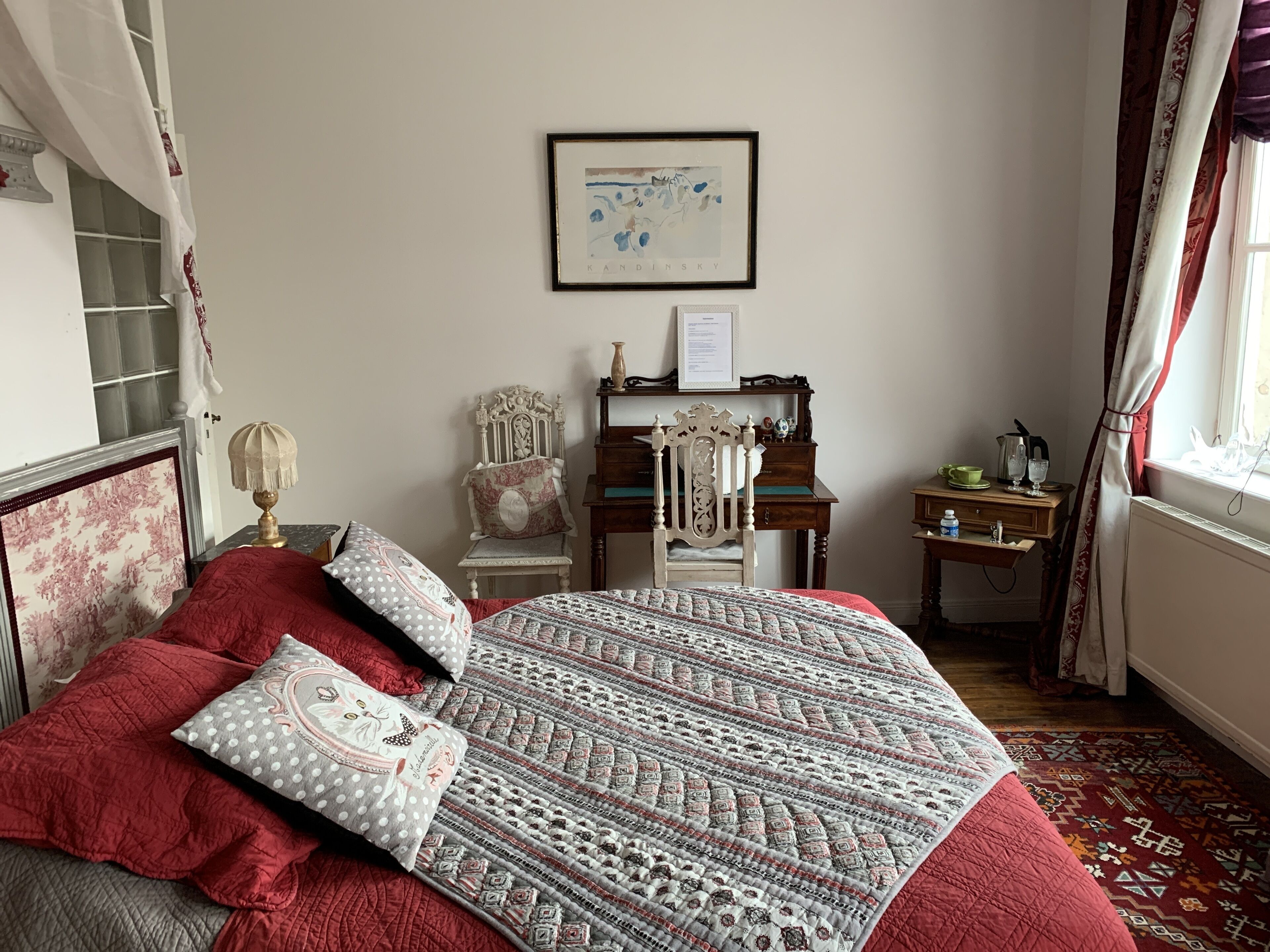 A bedroom with a large bed, a wooden dresser, a chair, a framed picture on the wall, and a window with curtains.