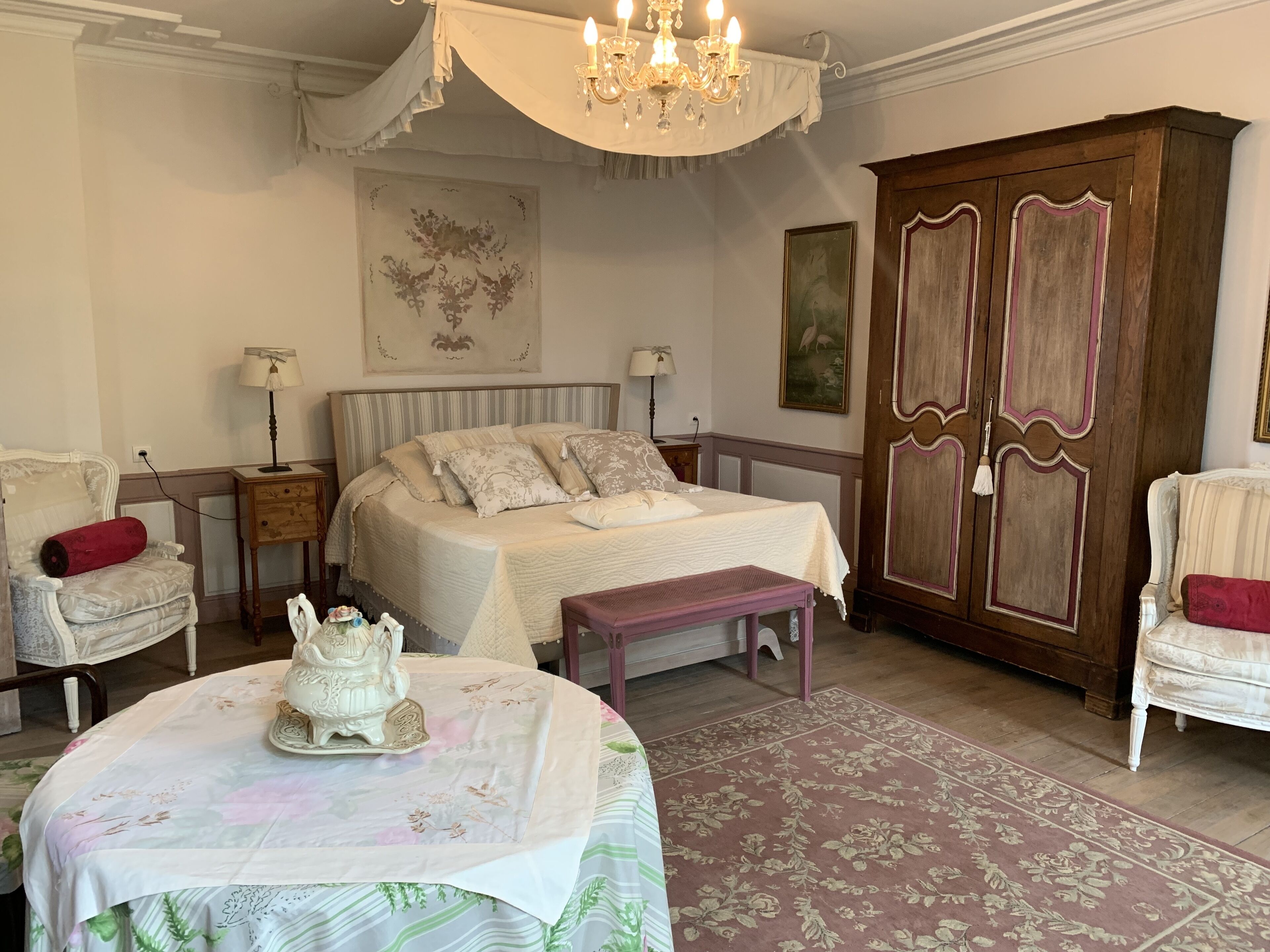 Ein Schlafzimmer mit einem großen Bett, einem Nachttisch, einer Kommode, einem Sessel, einem Tisch mit einem dekorativen Gegenstand und einer Chandelier.