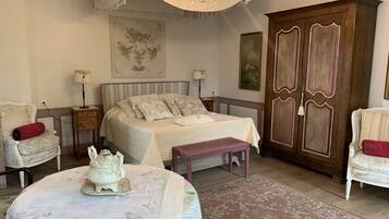Ein Schlafzimmer mit einem großen Bett, einem Nachttisch, einer Kommode, einem Sessel, einem Tisch mit einem dekorativen Gegenstand und einer Chandelier.