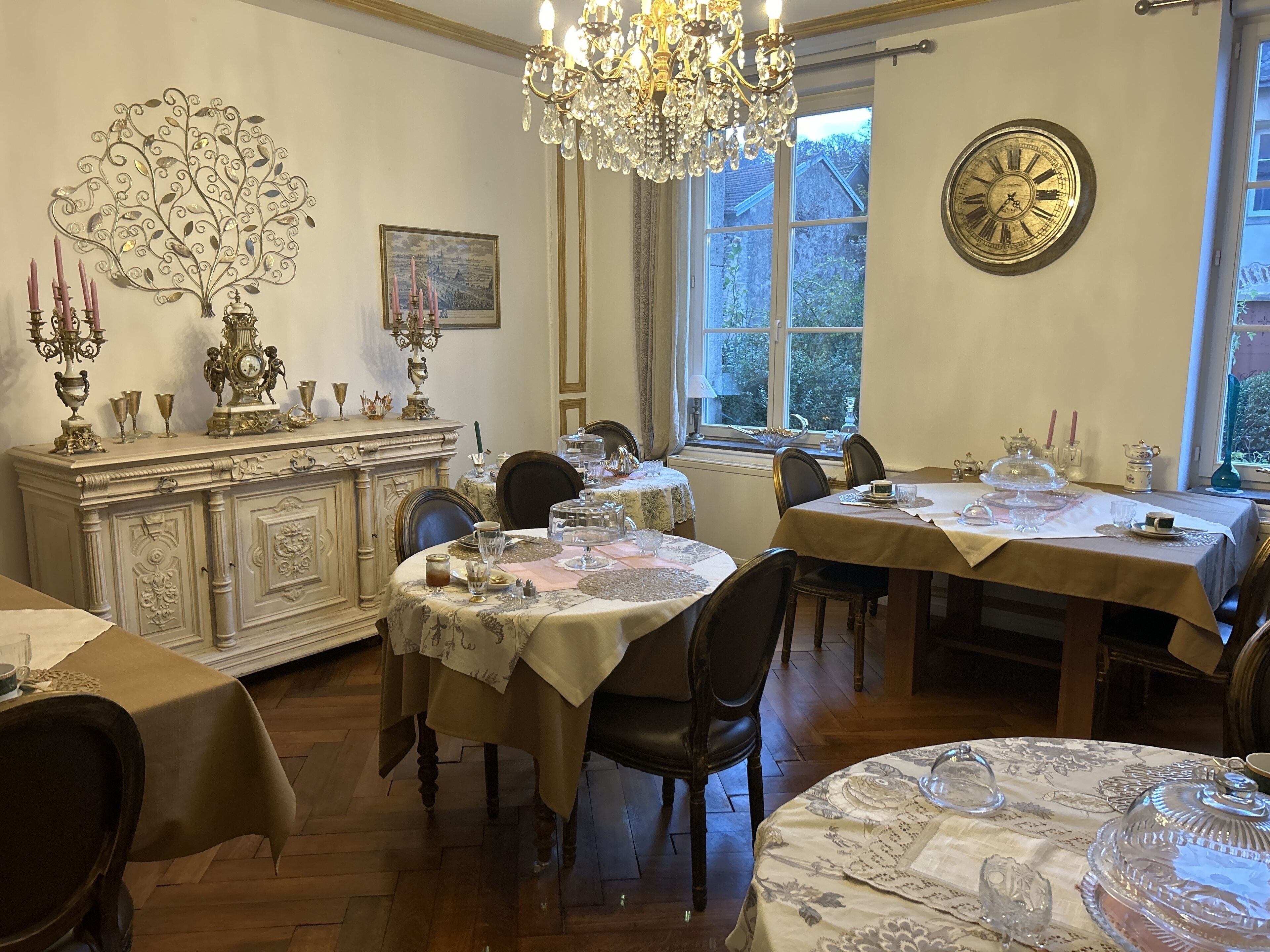 A dining room with ornate chandeliers, a large wall clock, and a view of the outdoors through a window.