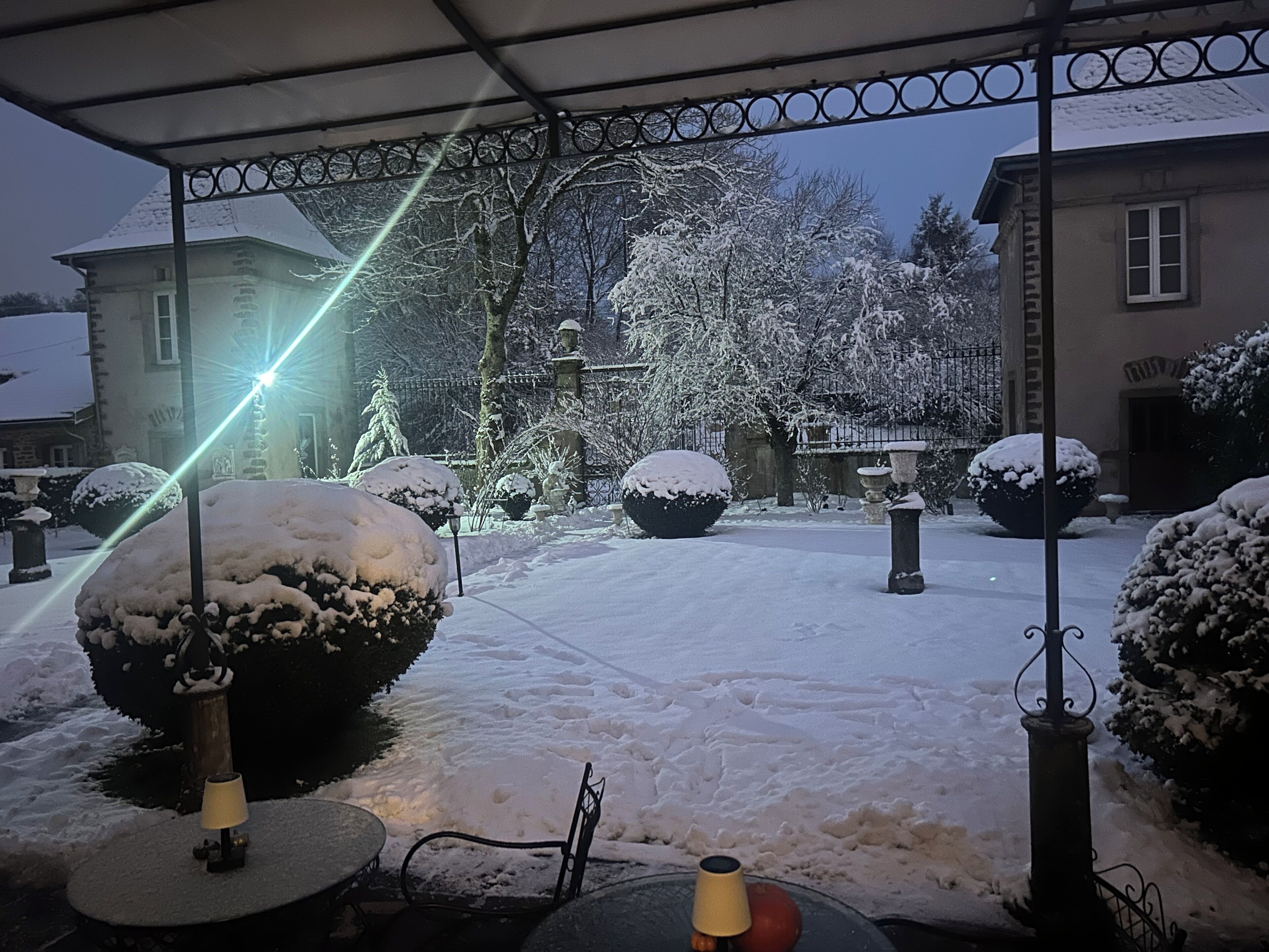 Un patio cubierto de nieve con un área de estar techada, árboles nevados y un edificio al fondo.