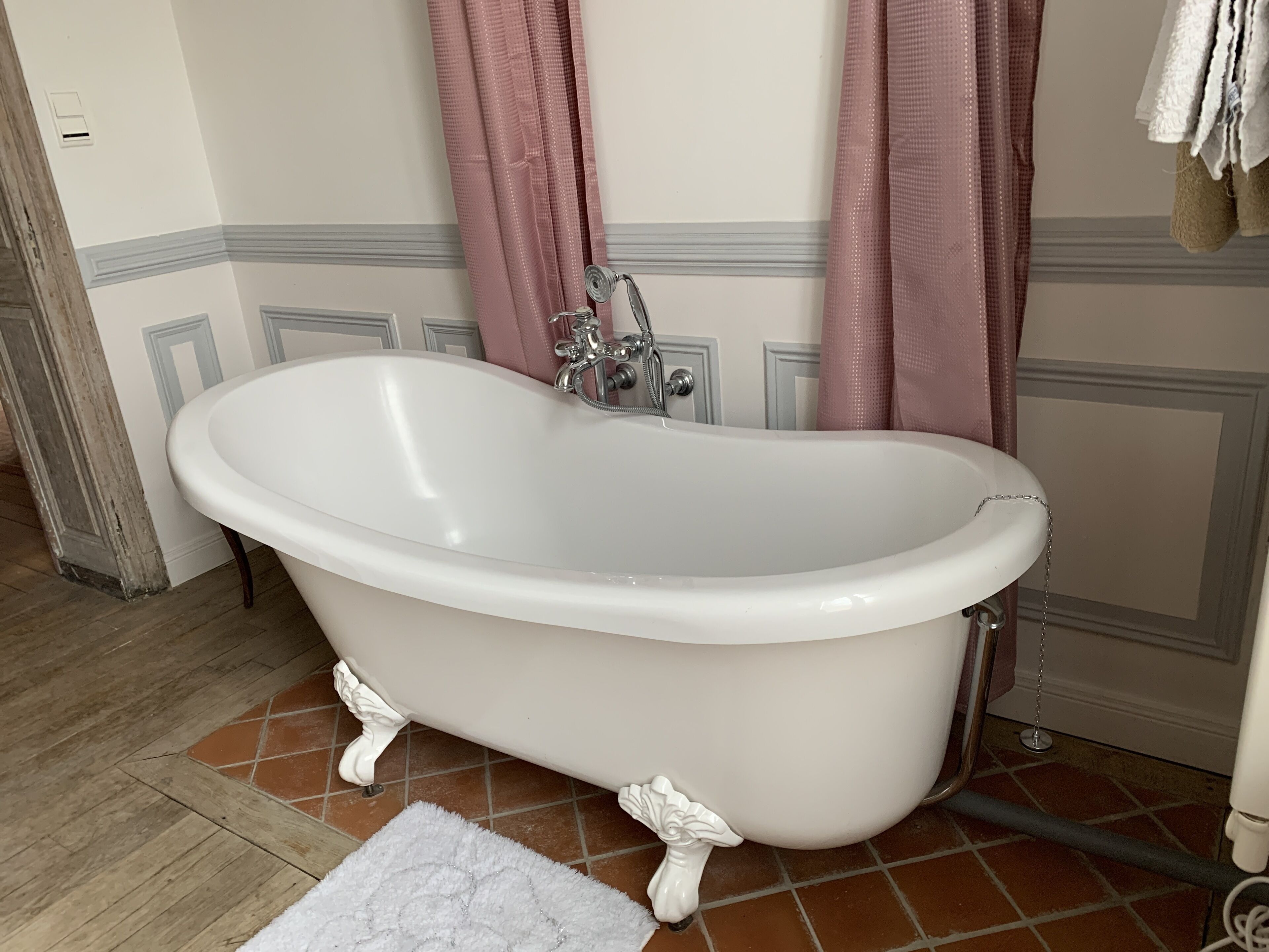 Une baignoire blanche, autoportante, sur pieds en forme de griffes, un rideau de douche et un tapis blanc dans une salle de bain dont le sol est carrelé et qui possède une porte avec un cadre en bois.