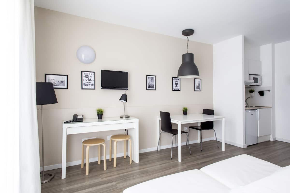 Een moderne, minimalistische kamer met een wit bureau, twee houten krukken, een zwarte stoel, een flatscreen televisie, een hanglamp en een kleine kitchenette.
