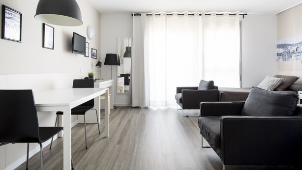 Een moderne woonkamer met een witte eettafel, zwarte stoelen, een grijze bank en een grote, zwarte hanglamp.