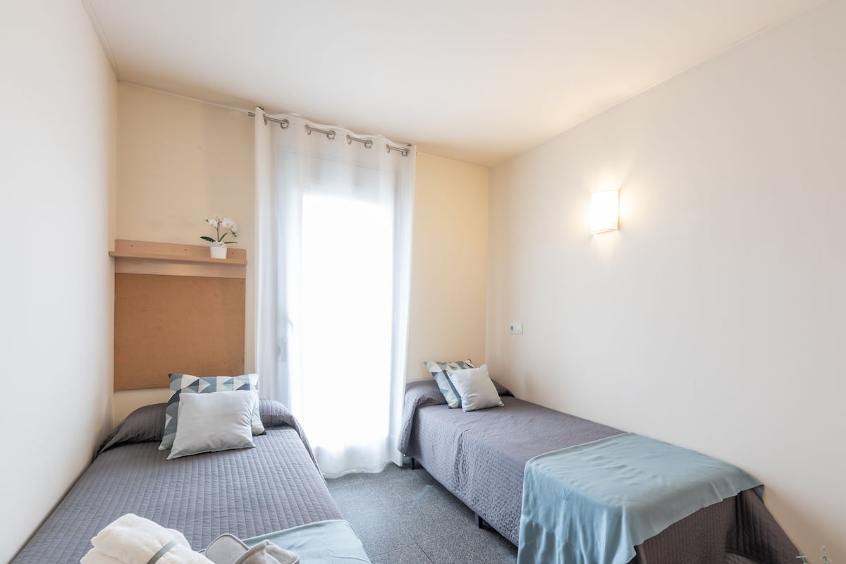 Een kleine, schone kamer met twee eenpersoonsbedden, een raam met gordijnen, een wandplank met een plant en een wandlamp.