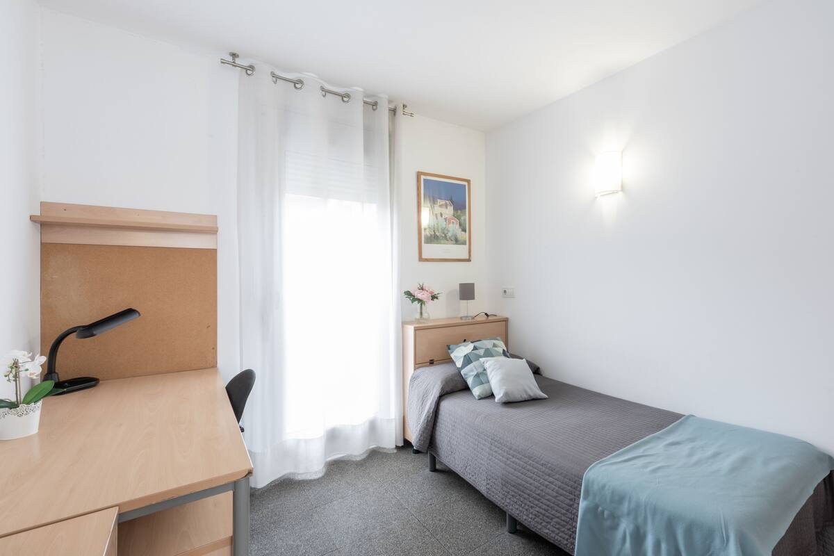 Een slaapkamer met een bureau, een bed, een nachtkastje, een lamp en een lijst met een afbeelding aan de muur.