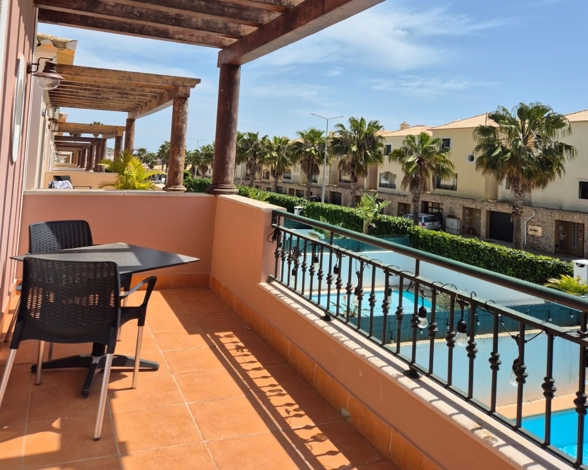 A balcony with a table and chairs, overlooking a swimming pool and palm trees.