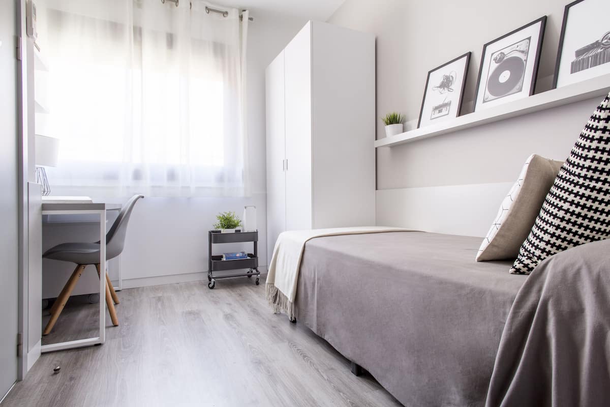 A modern bedroom with a bed, a desk, and a chair. There is a window with curtains, a shelf with framed pictures, and a small plant on the shelf.