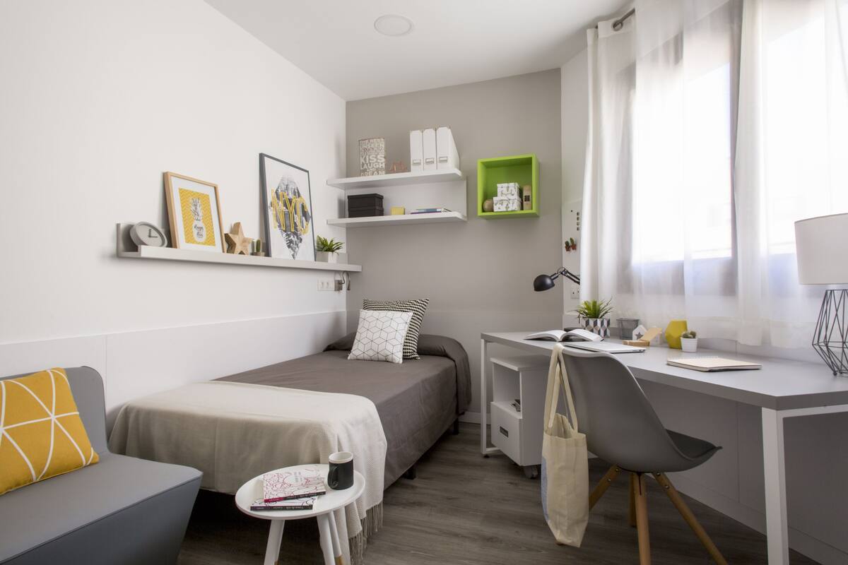 A modern, well-lit bedroom with a bed, bedside table, and a desk with a chair. There are shelves above the bed and a small table with a cup and a book.