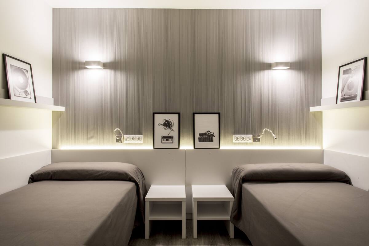 A modern hotel room with two beds, a central table, and wall-mounted lighting.