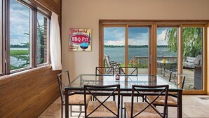 A dining area with a glass table and chairs, a sign reading "Backyard BBQ FIT," and a view of a body of water through large windows.