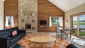 A spacious living room with a stone fireplace, a black leather sofa, a wooden coffee table, and a dining area with a view of a lake.
