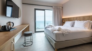 A modern hotel room with a large bed, a flat-screen TV, a wooden desk, and a view of the sea through the sliding glass door. - White Olive Elite Rethymno - All Inclusive (Rethymno)
