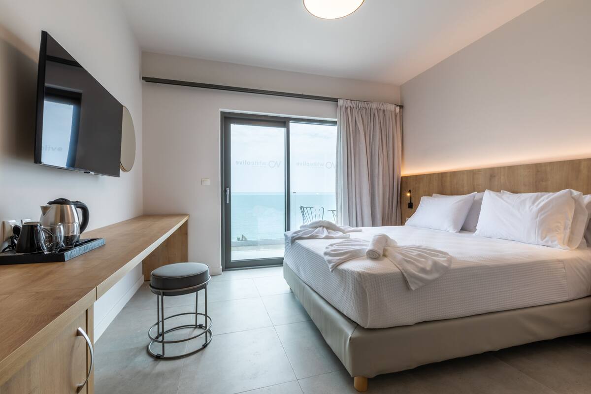 A modern hotel room with a large bed, a flat-screen TV, a wooden desk, and a view of the sea through the sliding glass door.