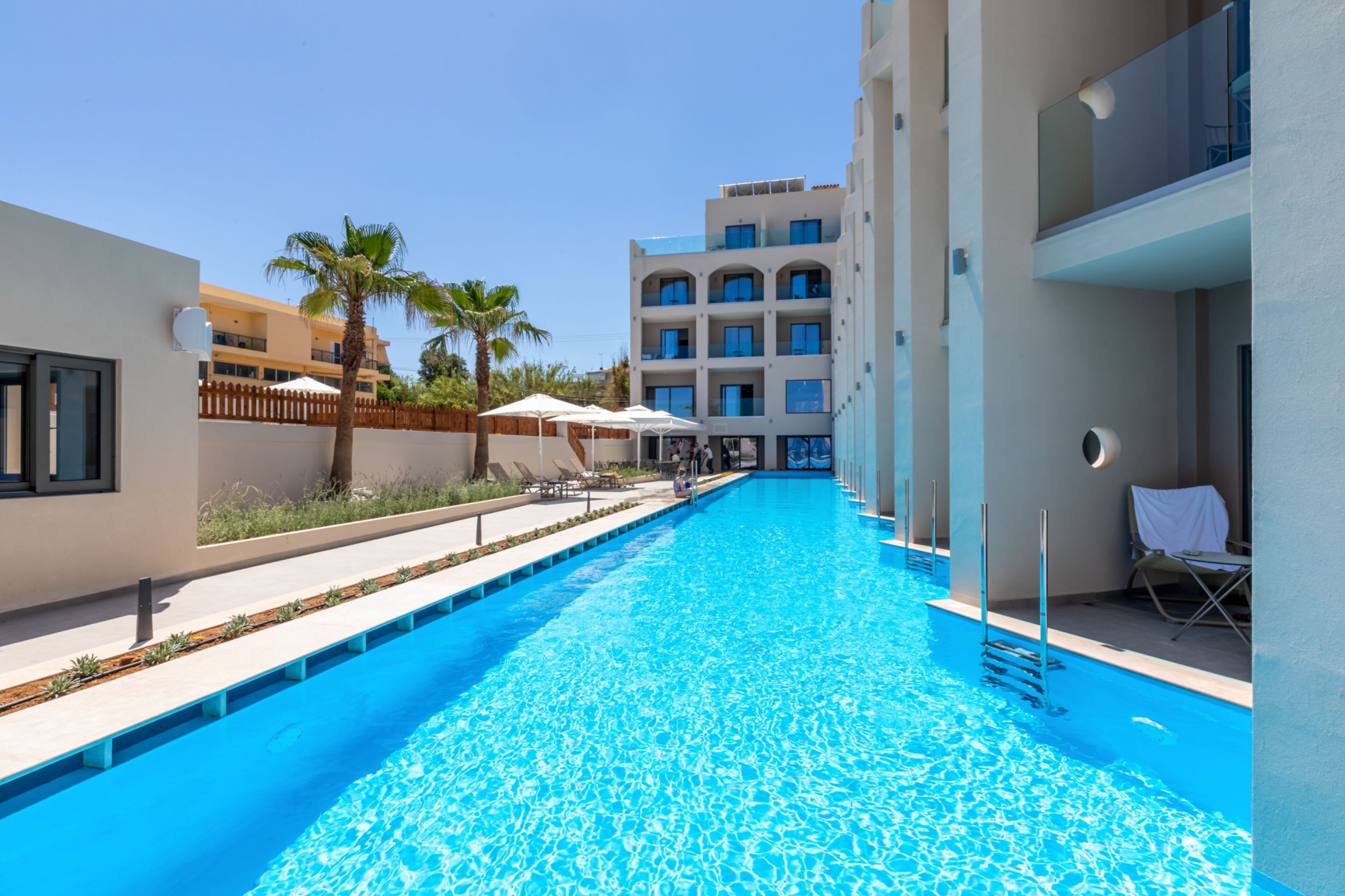 A swimming pool with clear blue water, surrounded by modern buildings, palm trees, and outdoor seating areas.