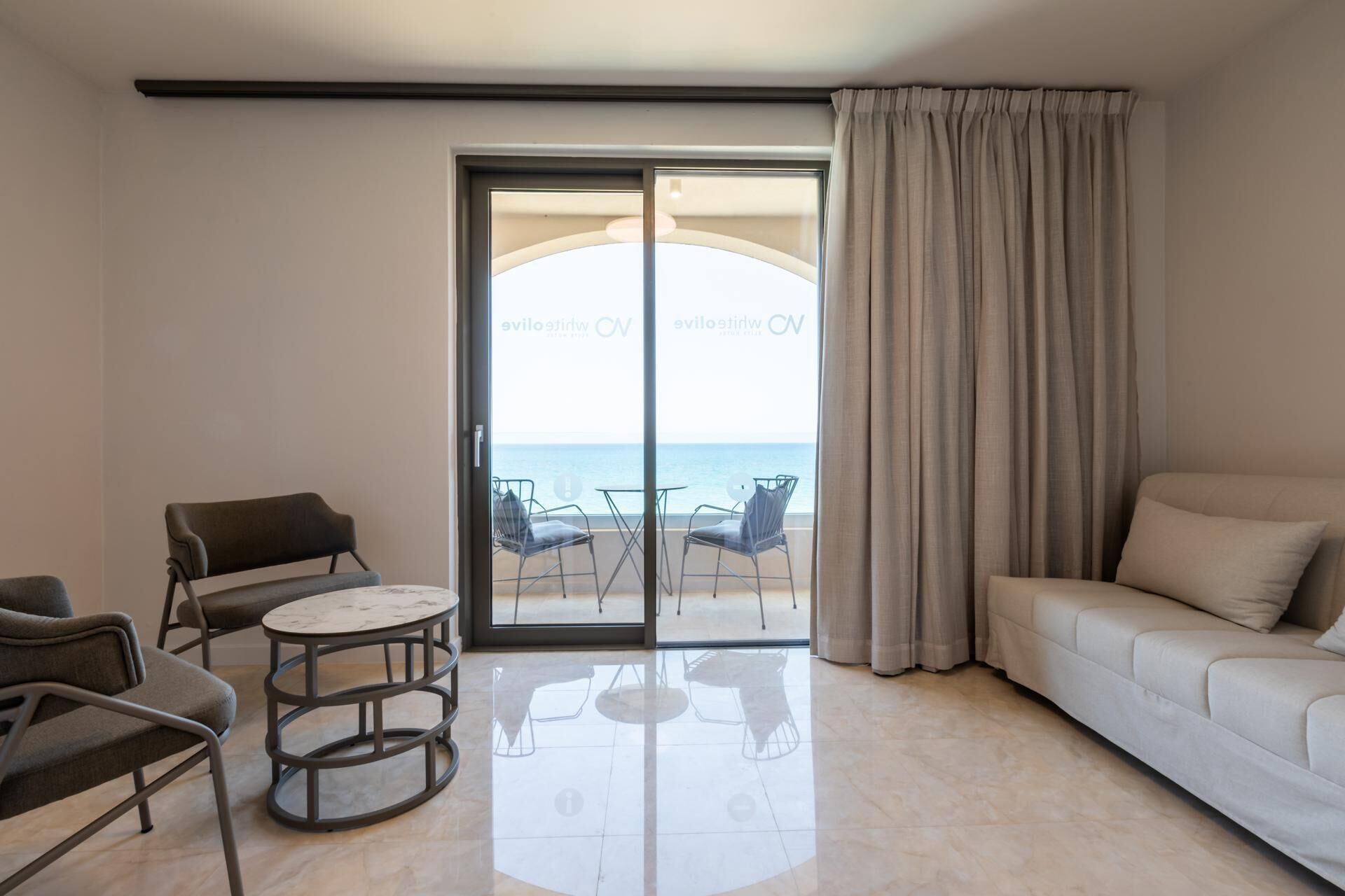 A hotel room with a sofa, a dining table with chairs, and a view of the sea through a glass door.