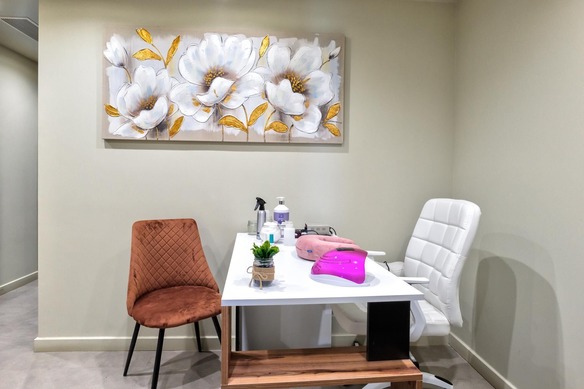 Una stanza con un tavolo bianco, due sedie, una poltrona massaggiante rosa e un grande quadro floreale appeso al muro.