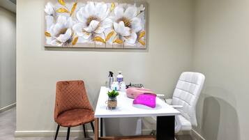 Una stanza con un tavolo bianco, due sedie, una poltrona massaggiante rosa e un grande quadro floreale appeso al muro.