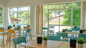 Un comedor luminoso con varias mesas y sillas, ventanas amplias con vista a árboles y edificios, y plantas en macetas sobre las mesas.