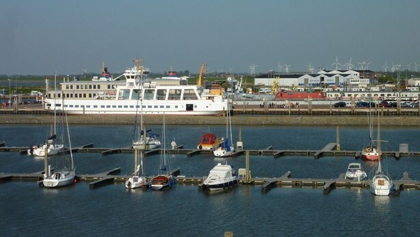 A marina with multiple boats docked, a large white ferry, and wind turbines in the background.