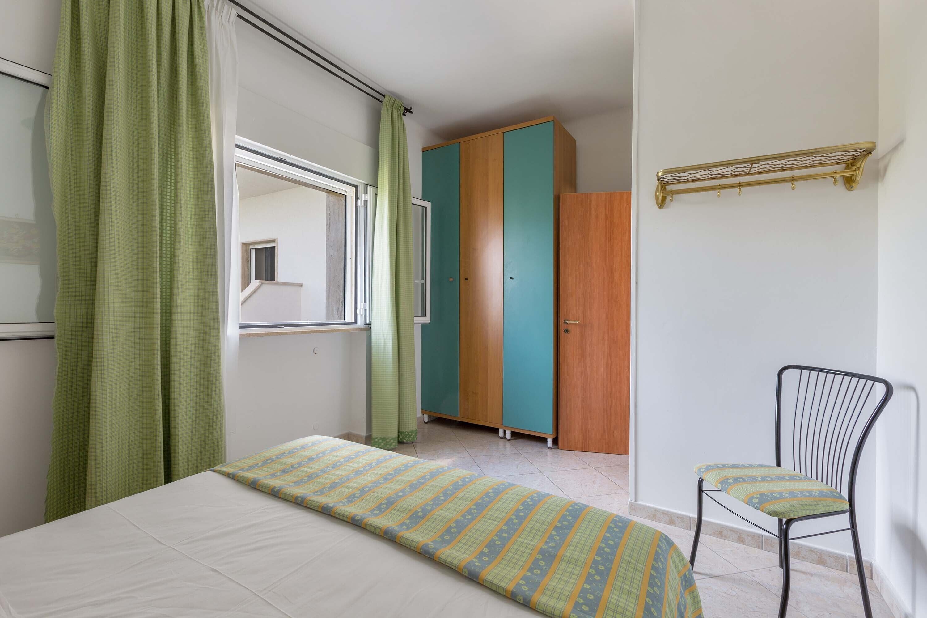 A bedroom with a bed, a chair, a wardrobe, and a window with curtains.