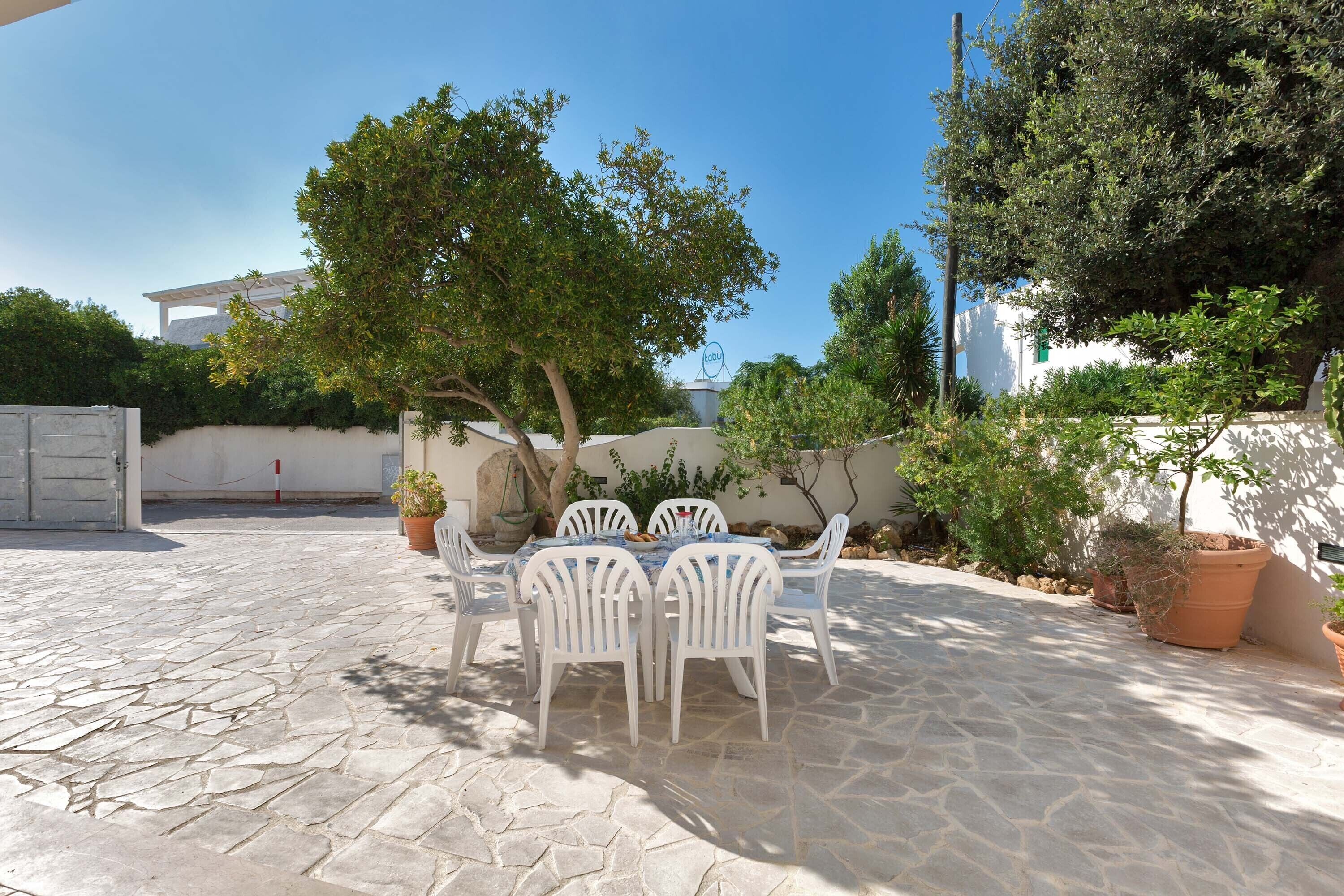 A patio with a stone floor, white plastic chairs, and a table. There are potted plants and a tree providing shade.