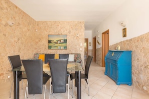 A dining area with a glass table and black chairs, a sofa with a floral cushion, and a blue cabinet against a stone wall.
