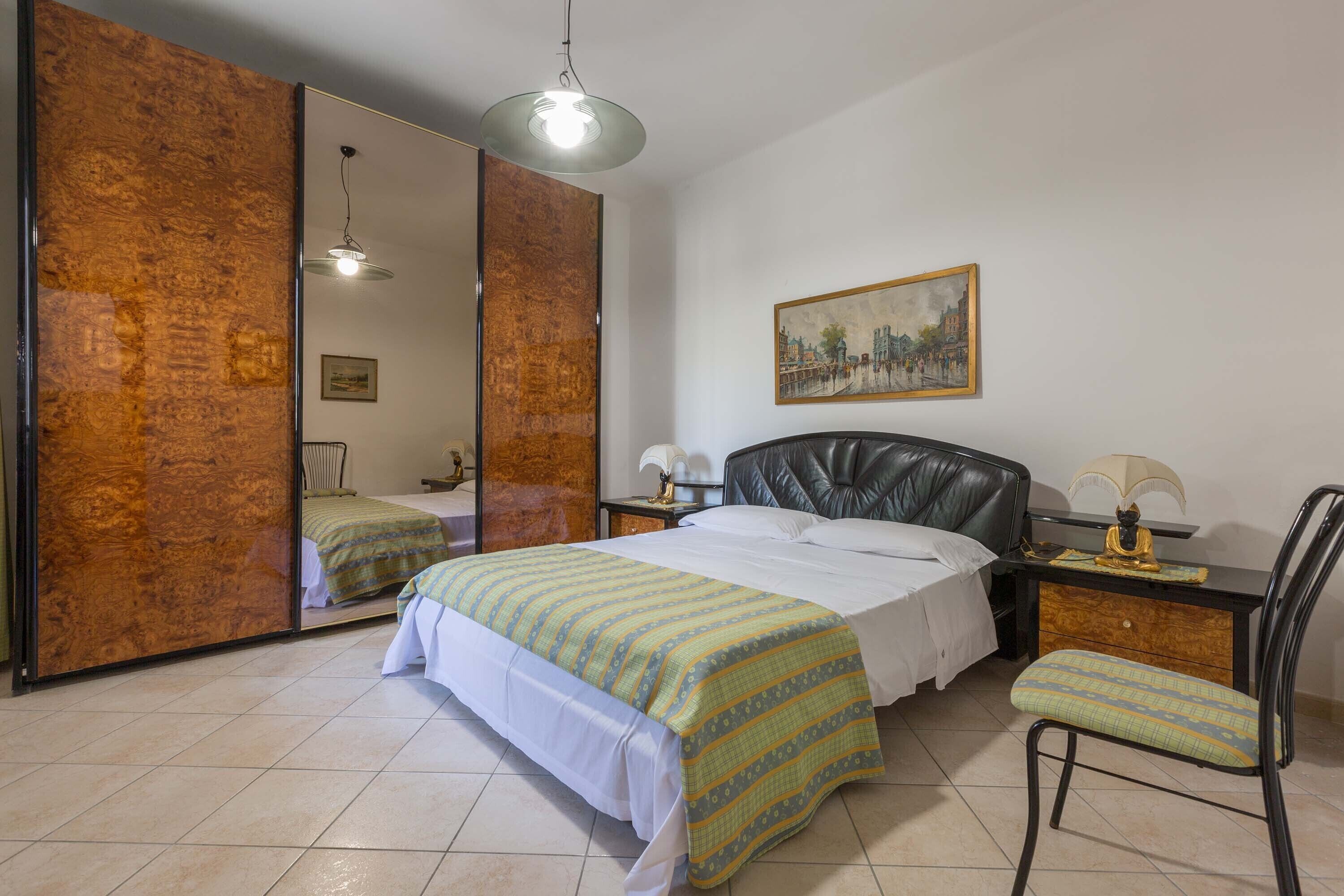 A bedroom with a bed, a chair, a desk, a mirror, and a painting on the wall.