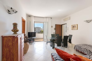 A modern living room with a sofa, a TV, a dining table with chairs, and a balcony with a view.