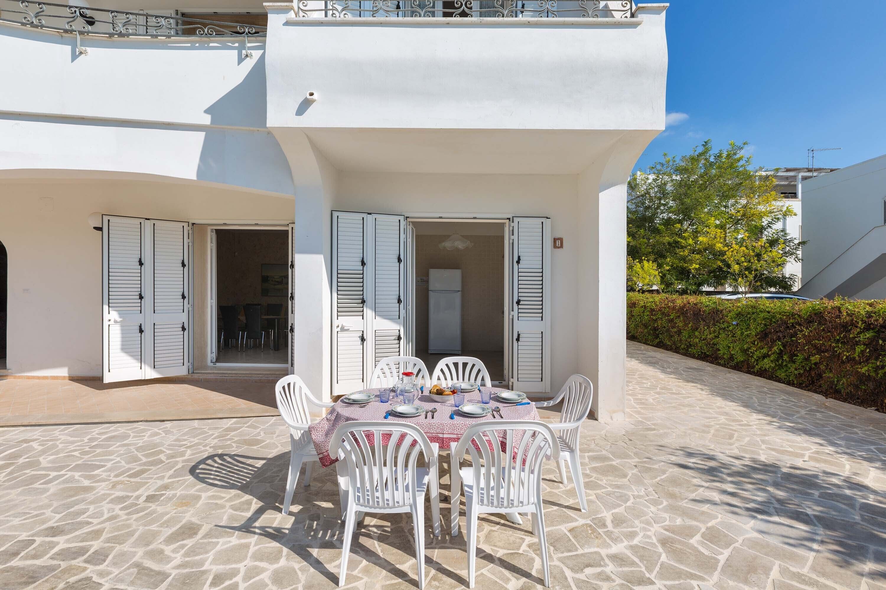 Un espace de restauration en plein air avec une table dressée pour quatre personnes, entourée de chaises blanches, sous un bâtiment blanc doté de volets.