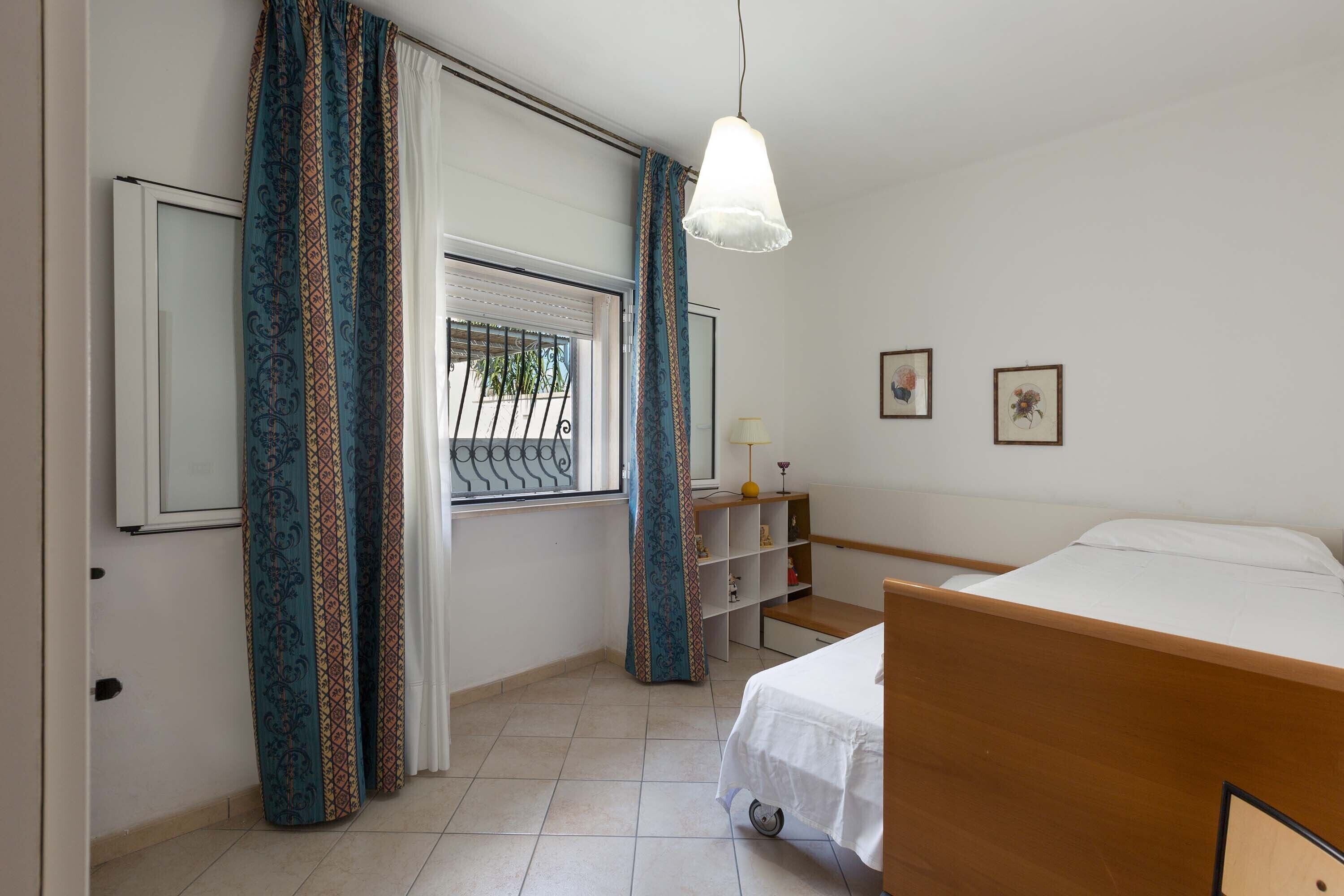 A bedroom with a bed, a window with curtains, a bedside table with a lamp, and a shelf with decorative items.