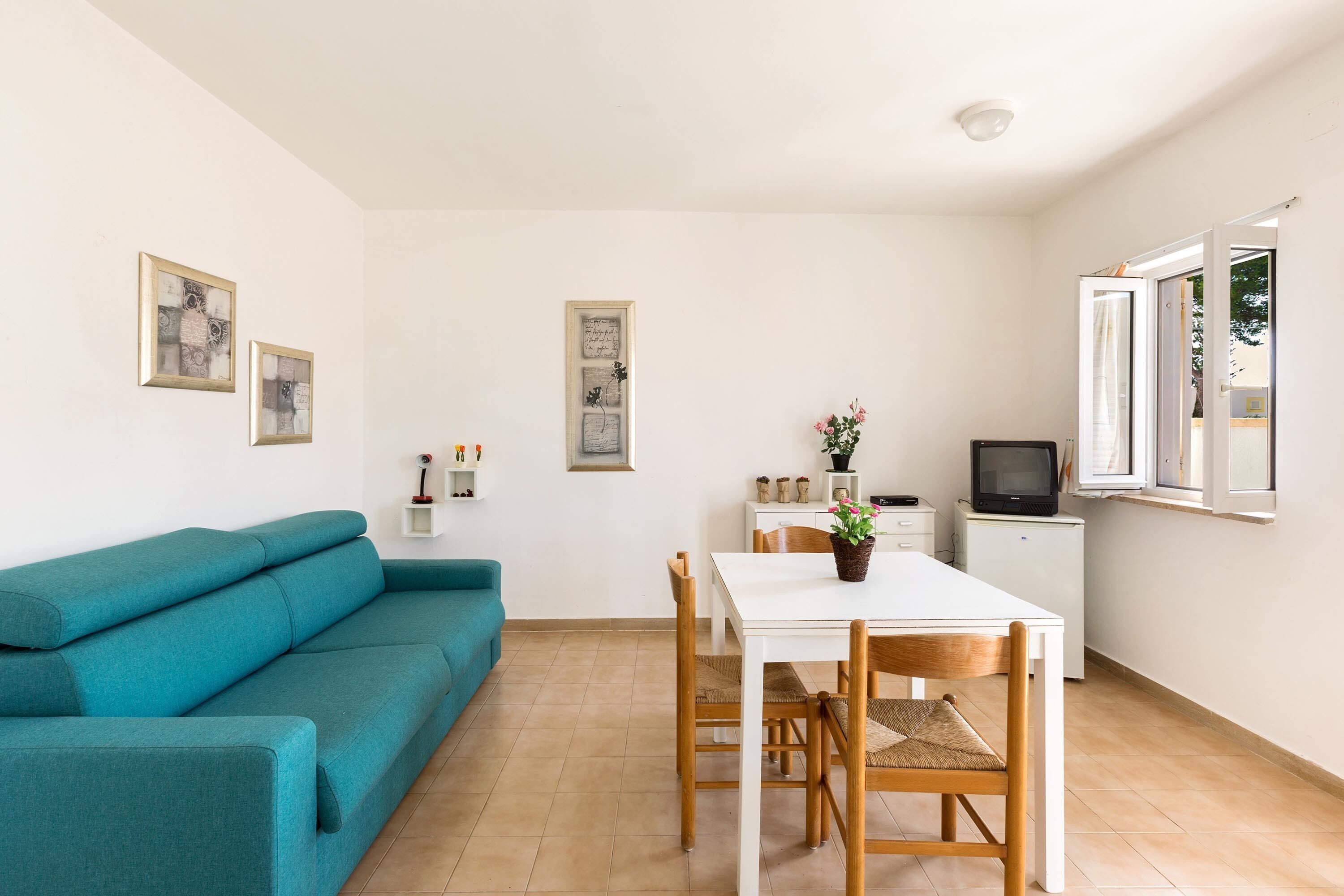 A modern living room with a teal sofa, a white dining table with chairs, a small TV on a stand, and artwork on the walls.