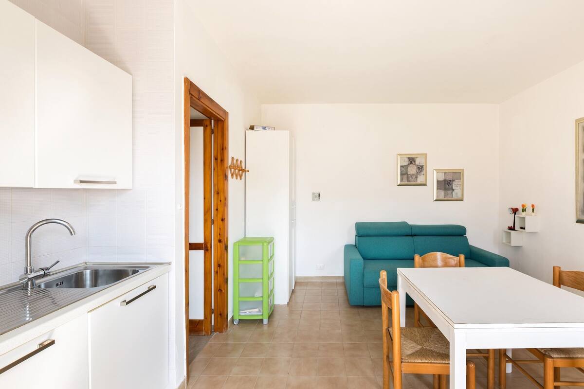 A compact kitchen with a sink, cabinets, and a green storage cart. A teal sofa and a white table with chairs are in the living area.
