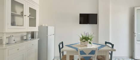 A compact kitchen with white cabinetry, a built-in refrigerator, a dining table with chairs, and a mounted flat-screen TV.