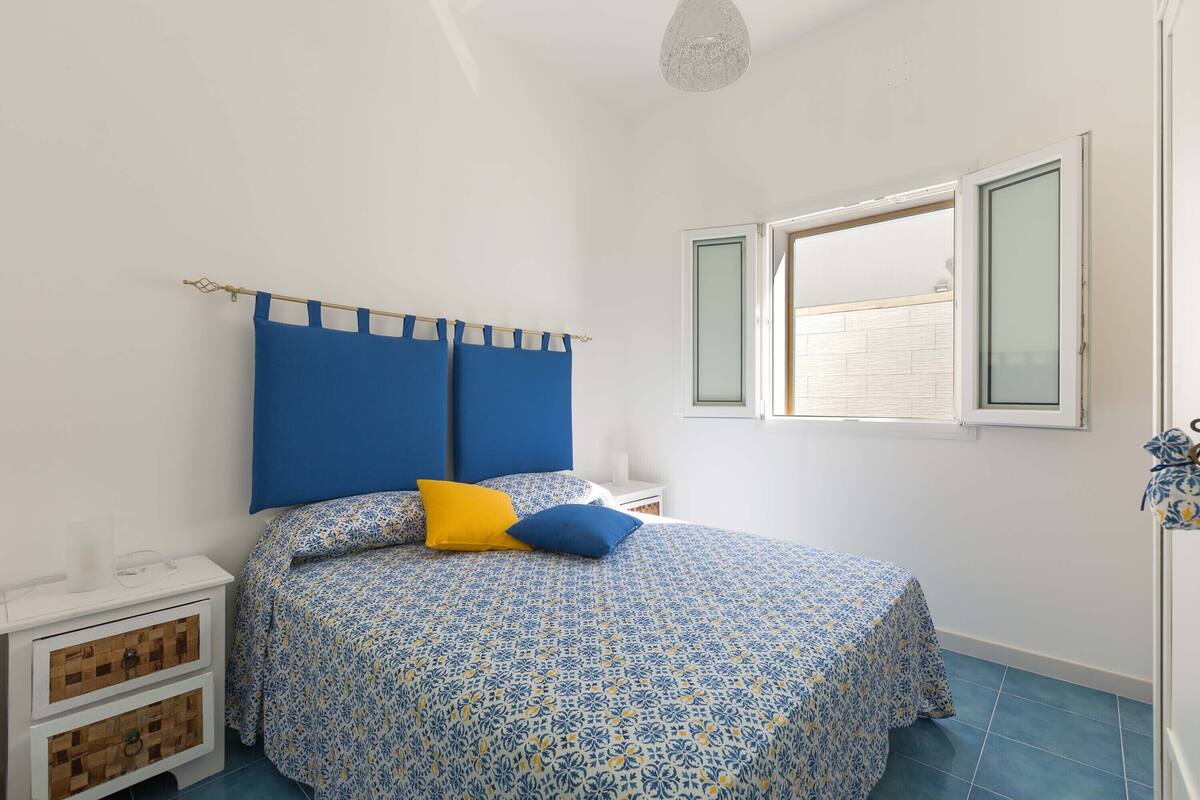 Un dormitorio con cama, mesita de noche, ventana y puerta.