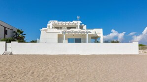 Un bâtiment moderne situé en bord de mer, avec une façade blanche, un espace extérieur couvert et un ciel d’un bleu limpide.