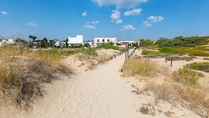 A sandy pathway leading to a white building with arches, surrounded by greenery and a clear blue sky. - Villa Oasi Marina (Maruggio)