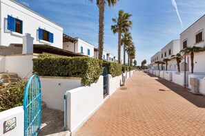 Una calle empedrada y estrecha flanqueada por edificios blancos, palmeras y un portón azul.