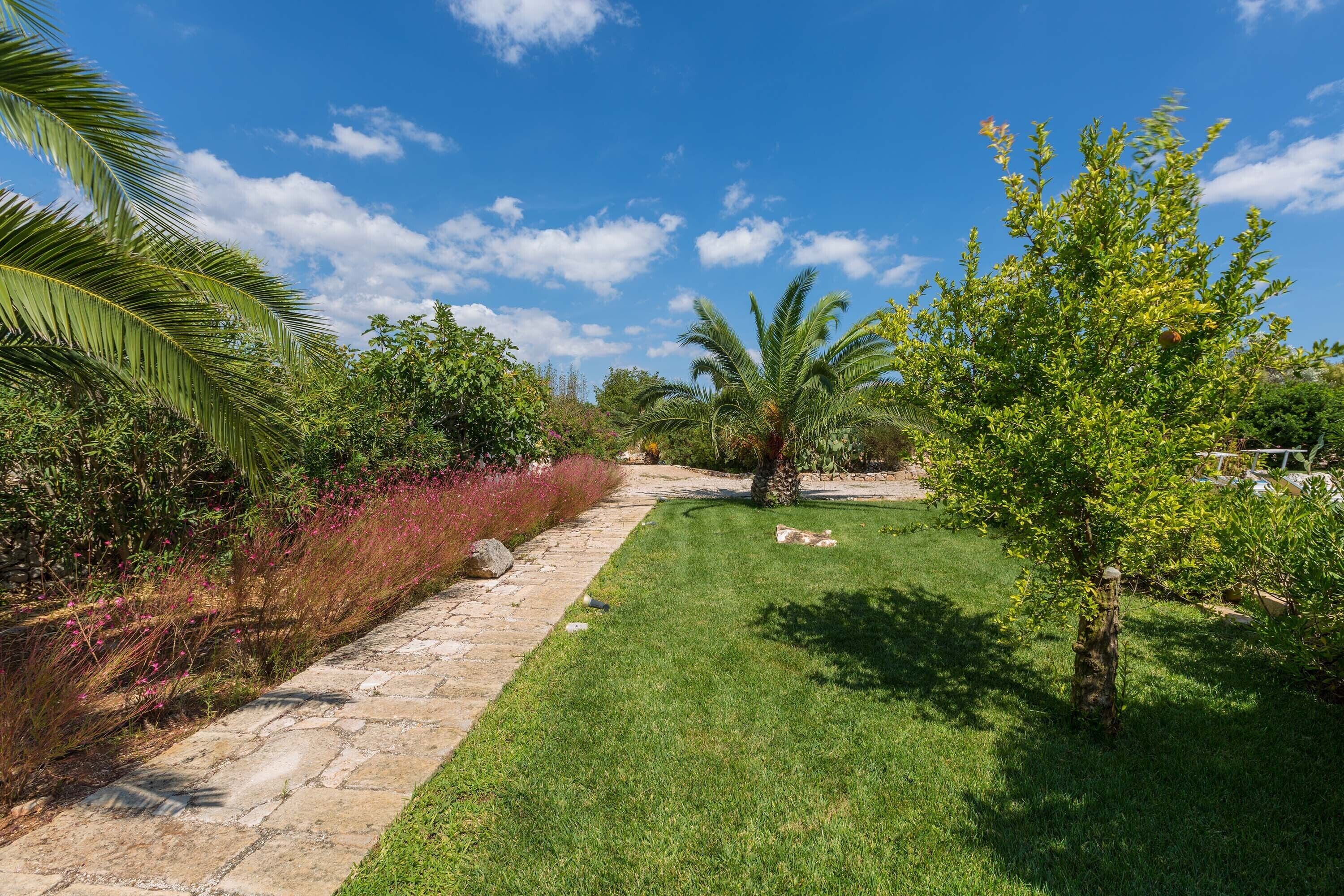 A well-maintained garden with a stone pathway, lush green grass, and various plants including palm trees and flowering shrubs.
