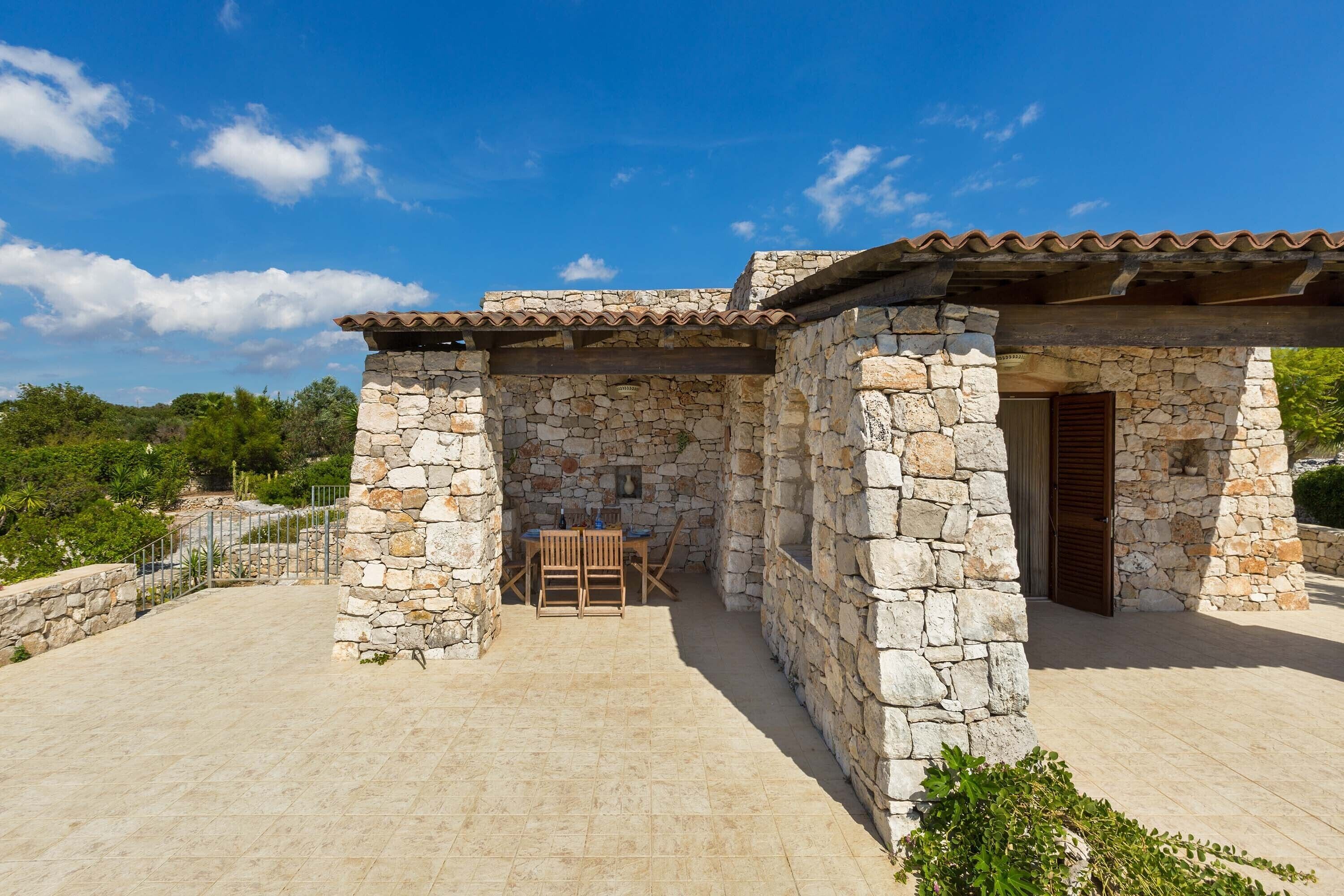 A stone building with a tiled roof, a patio with a table and chairs, and a stone wall.