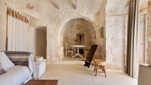 A rustic room with a stone wall, a wooden table with chairs, a sofa, and a small stool. - Trullo Giamar (Ostuni)