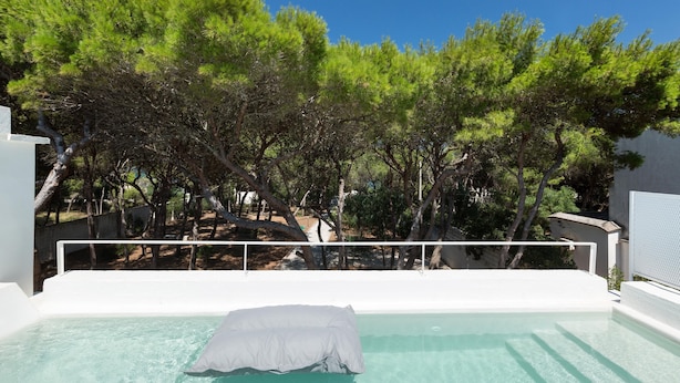 A clear swimming pool with a white edge, surrounded by trees and a white wall. - Masseria Villa (Melendugno)