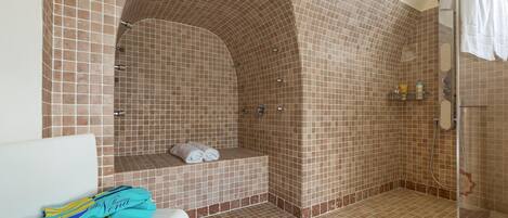 Une salle de bain avec une douche carrelée, un banc avec des serviettes et un lavabo blanc.