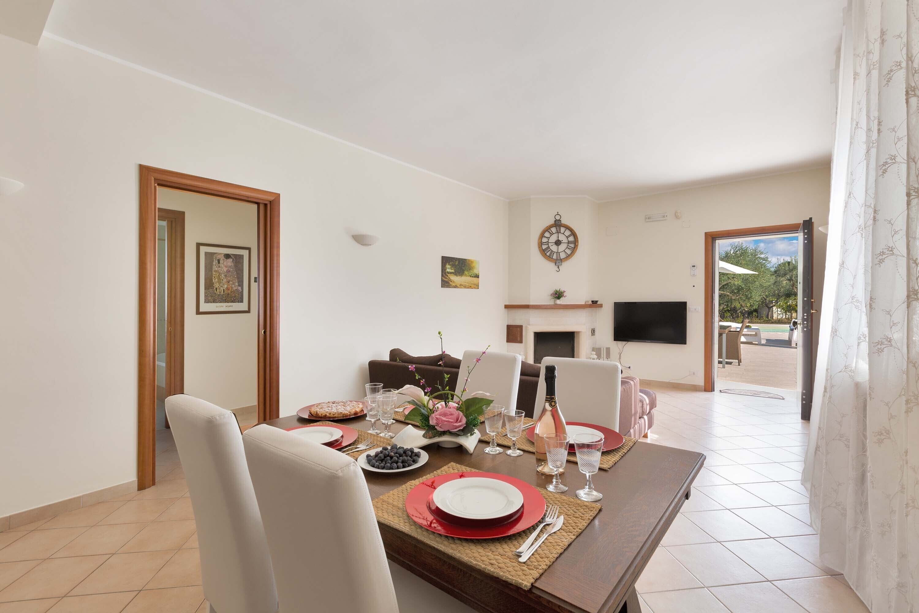 A dining area with a wooden table set for four, white chairs, and a television mounted on the wall.